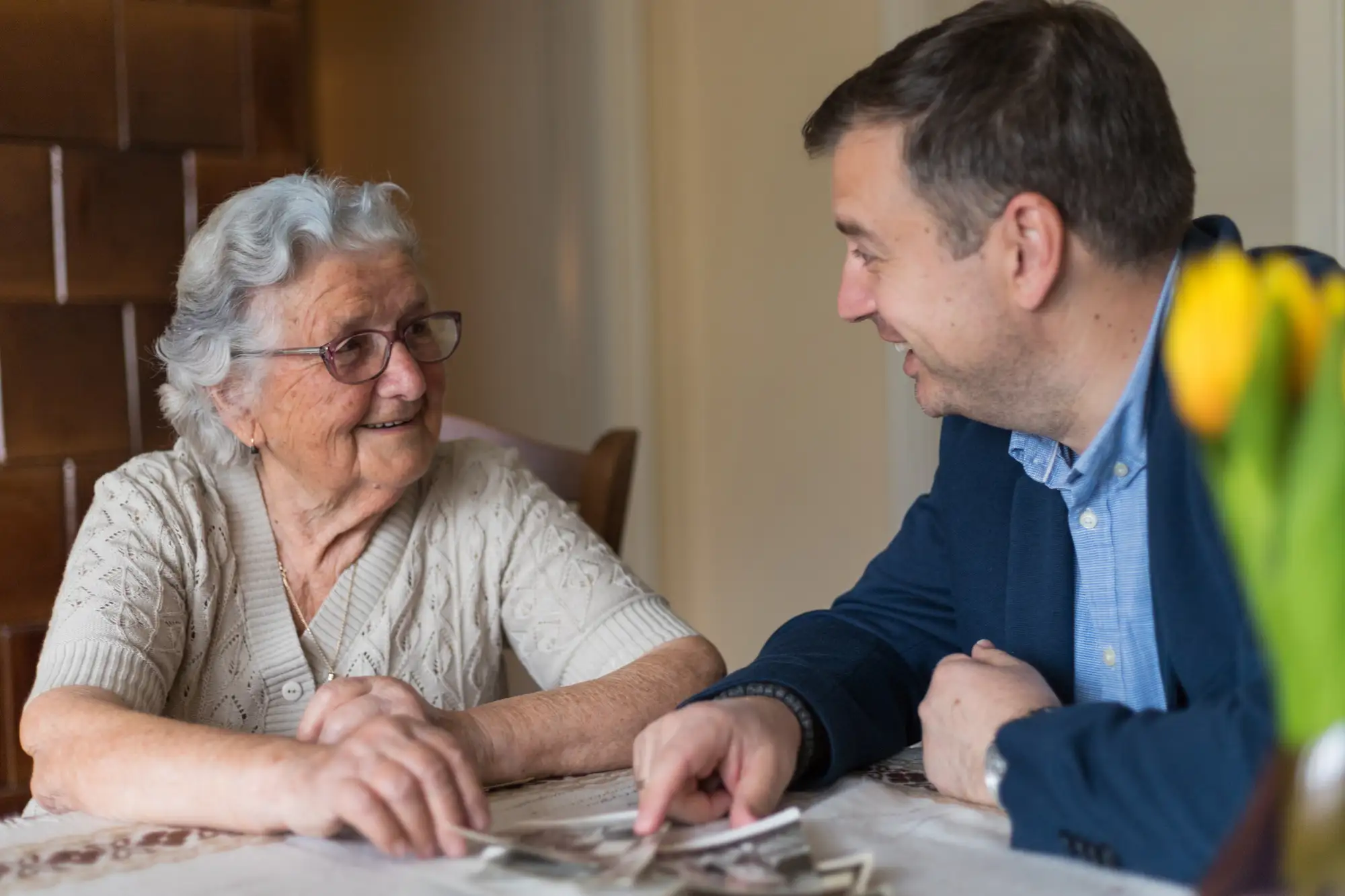 Old woman with gray hair, glasses and a middle-aged man in a jacket are sitting at the table and looking at the photos in the old family house.